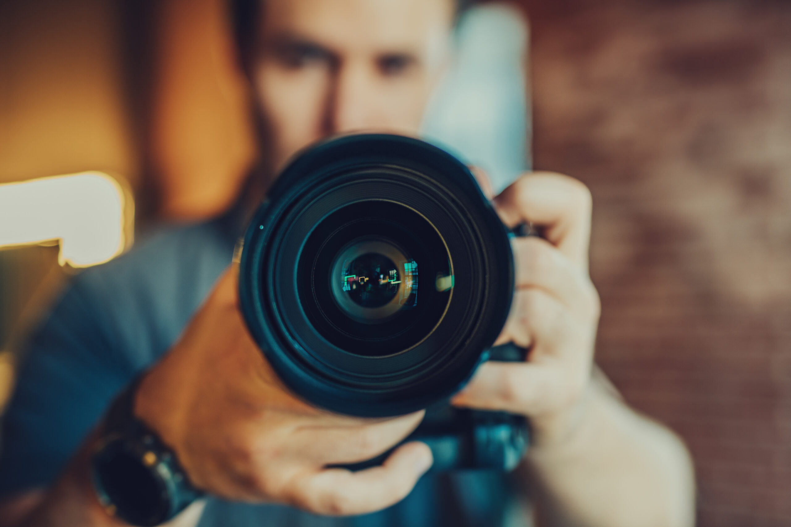 image of camera lens, male photographer in blue shirt holding the camera in background